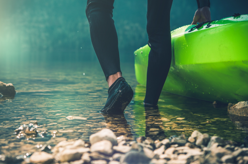 Best water shoes for beach and river activities worn in shallow rocky water beside a kayak.