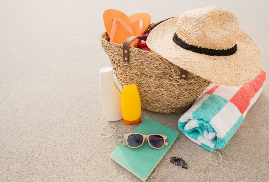 Beach day packing list with a beach bag, towel, sunscreen, hat, sunglasses, sandals, and book on sand.